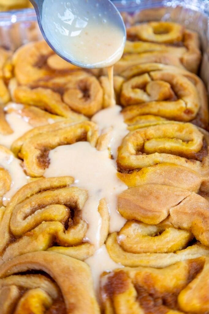 Cream cheese frosting being poured over warm pumpkin cinnamon rolls in a baking pan, showing the frosting melting into the spirals.