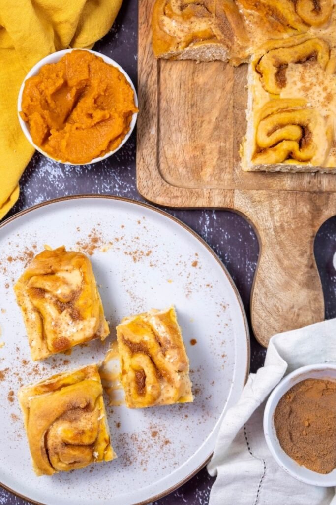 Soft pumpkin cinnamon rolls with cream cheese frosting served on a plate, showing the swirled pumpkin filling and tender interior, with additional rolls and pumpkin purée in the background.