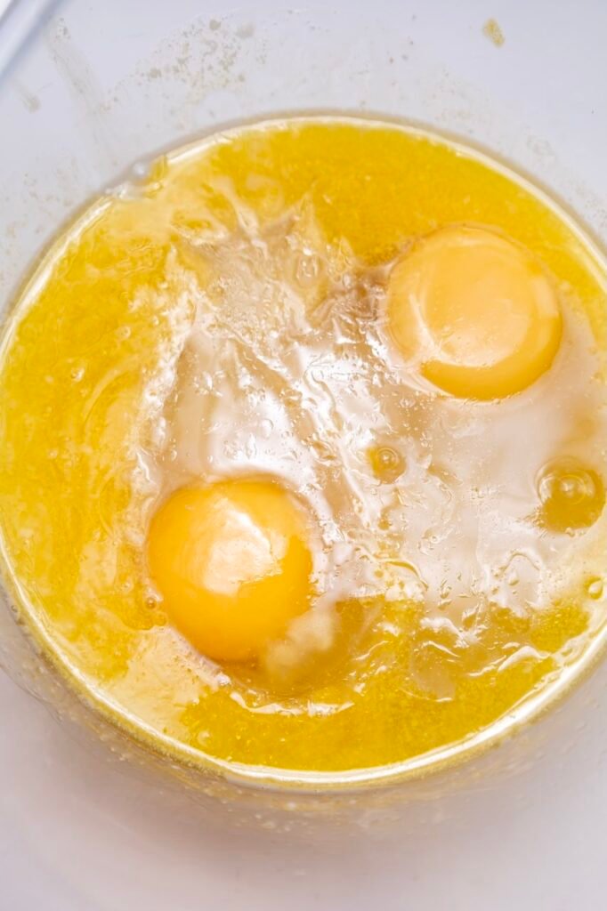 Melted butter and cracked eggs added to a bowl of warm milk and yeast, showing the mixture before being whisked together to start pumpkin cinnamon roll dough.