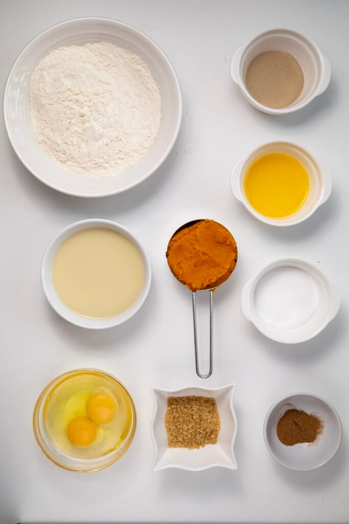 A flat lay of ingredients for pumpkin cinnamon rolls, including flour, yeast, warm milk, melted butter, pumpkin purée, eggs, sugar, brown sugar, and cinnamon arranged in white bowls on a light background.