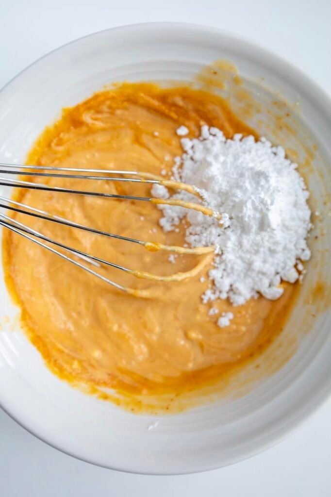 Powdered sugar being added to a bowl of pumpkin cream cheese frosting while whisking, showing the frosting thickening as it is mixed.