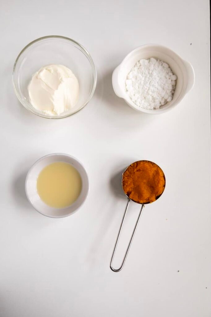 A flat lay of pumpkin cream cheese frosting ingredients, including softened cream cheese, powdered sugar, pumpkin purée in a measuring cup, and milk arranged on a light background.