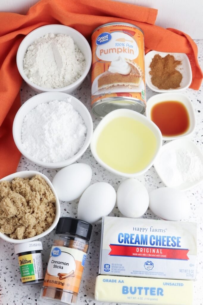 A flat lay of ingredients for pumpkin mini Bundt cakes arranged on a light countertop, including canned pumpkin purée, flour, brown sugar, eggs, oil, vanilla, pumpkin spice, cinnamon sticks, cream cheese, butter, powdered sugar, and food coloring.