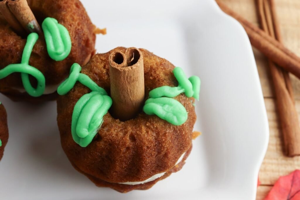 Close-up view of a pumpkin mini Bundt cake made from pumpkin spice cake with a visible layer of cream cheese frosting in the center. The cake is decorated with green frosting leaves and a cinnamon stick stem, sitting on a white plate with cinnamon sticks and fall decor softly visible in the background.