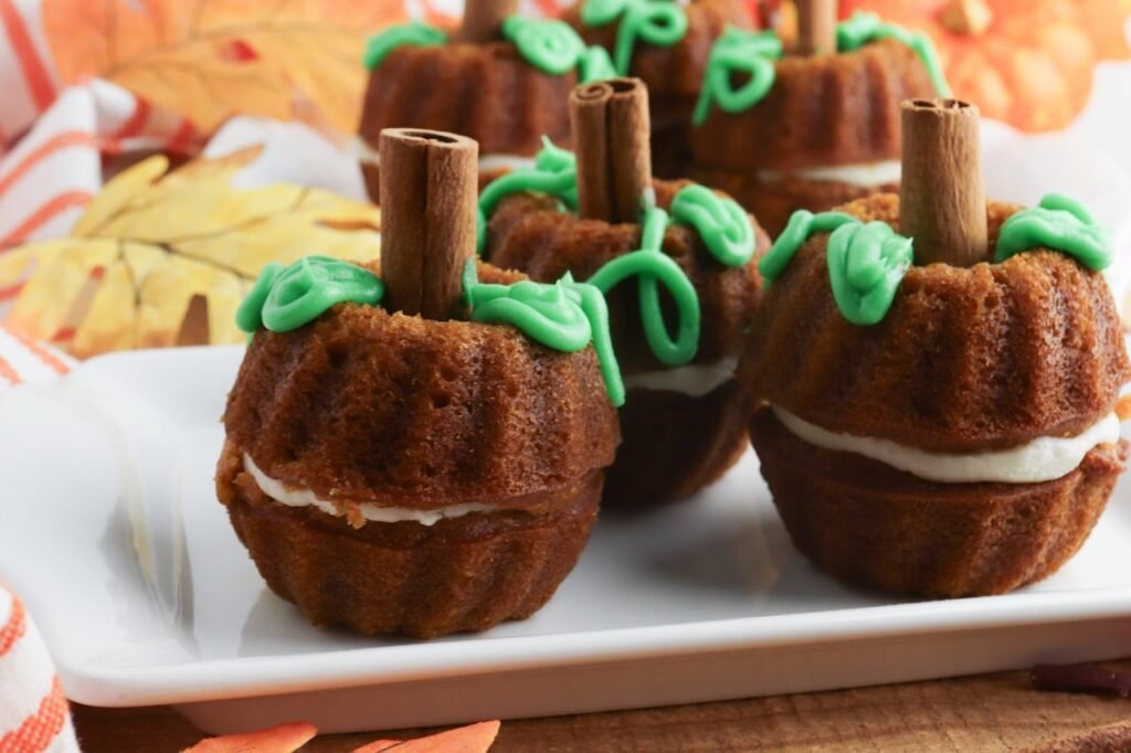 Close-up of pumpkin mini Bundt cakes made from moist pumpkin spice cake and filled with a layer of cream cheese frosting. Each mini cake is decorated with green frosting vines and a cinnamon stick stem, styled on a white serving tray with fall leaves and seasonal decor in the background.