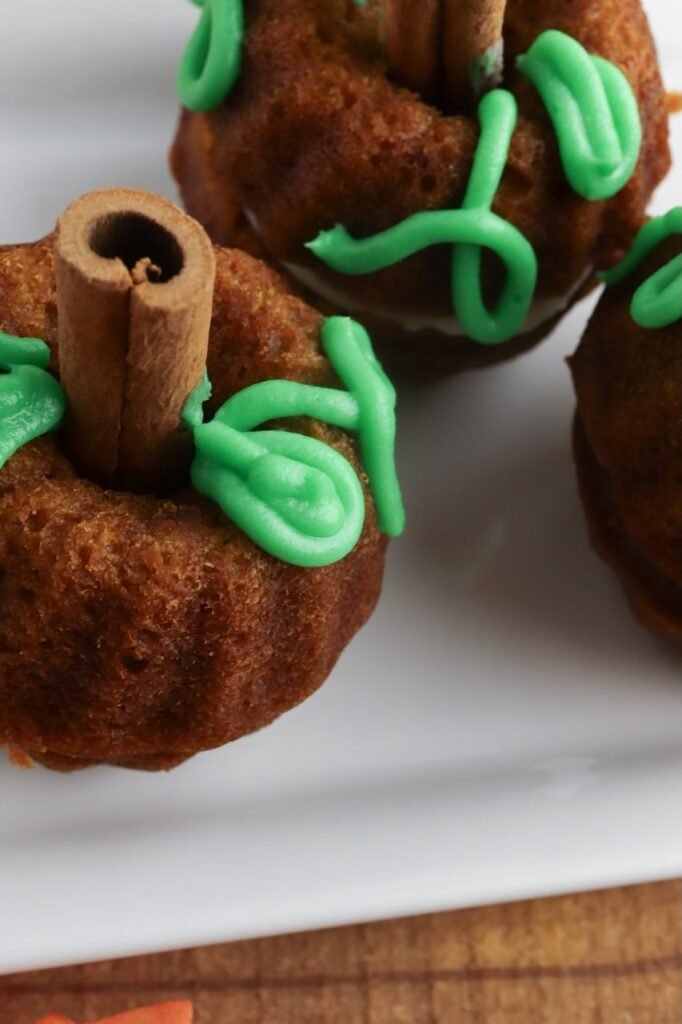 Close-up detail of pumpkin mini Bundt cakes made with pumpkin spice cake, showing the textured cake crumb, green frosting vines piped on top, and cinnamon stick stems. The cakes are arranged on a white plate with a wooden surface and soft fall decor visible in the background.