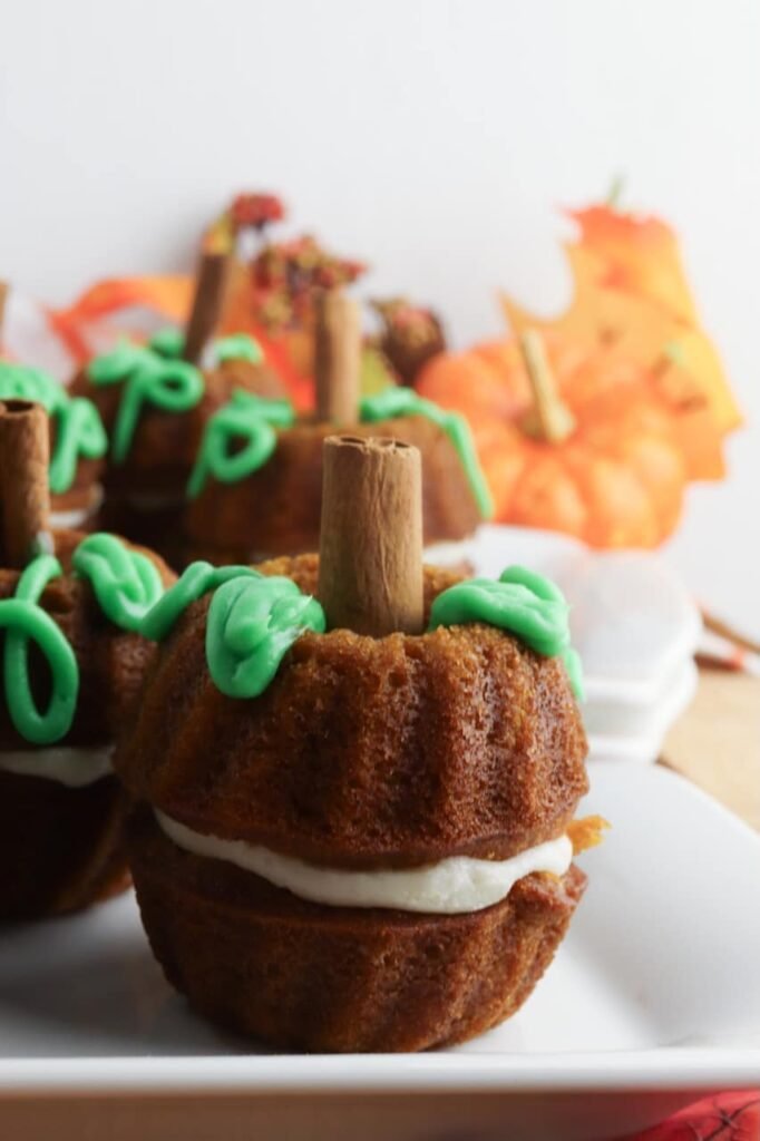 Close-up of a pumpkin mini Bundt cake made with moist pumpkin spice cake and a layer of cream cheese frosting in the center. The cake is decorated with green frosting vines and a cinnamon stick stem, styled on a white serving tray with soft fall decor and pumpkins blurred in the background.