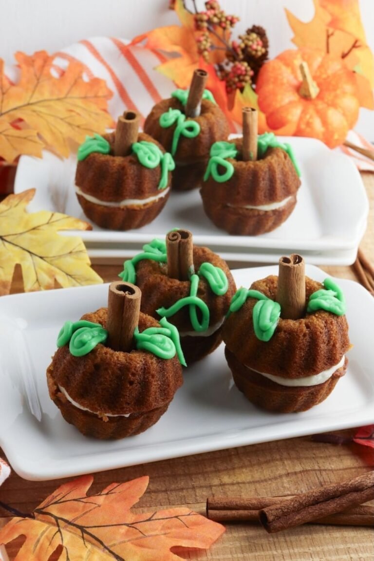 A serving platter filled with decorated pumpkin mini Bundt cakes made from pumpkin spice cake, layered with cream cheese frosting, and topped with green frosting vines and cinnamon stick stems. The cakes are styled to resemble small pumpkins and arranged on white plates with fall leaves and seasonal decor in the background.
