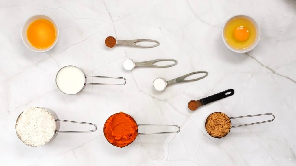 Overhead view of pumpkin pancake ingredients arranged on a light countertop, including measured flour, pumpkin purée, milk, brown sugar, cinnamon, nutmeg, baking powder, baking soda, and a cracked egg in a small bowl, prepared for mixing.