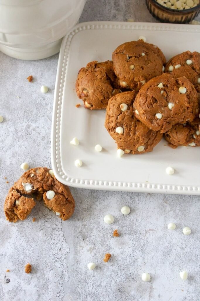 A stack of soft pumpkin spice cookies studded with white cream cheese-flavored chips sits on a white plate on a light gray surface. A broken cookie with a tender, cake-like crumb rests nearby, with a few chips and crumbs scattered around, showing the cookies’ thick texture and cozy fall-style bake.