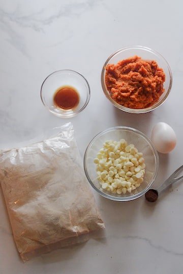 Overhead photo of pumpkin spice cookie ingredients on a light marble counter, including a bowl of pumpkin puree, a small bowl of vanilla extract, a bag of spice cake mix, a bowl of cream cheese-flavored baking chips, a whole egg, and a measuring spoon with ground spice.
