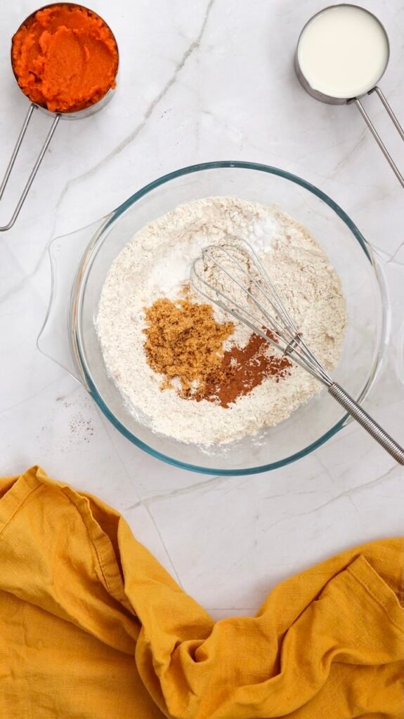 Overhead view of a glass mixing bowl with flour, brown sugar, cinnamon, nutmeg, baking powder, and baking soda being whisked together, showing the dry ingredients prepared for making pumpkin pancake batter.