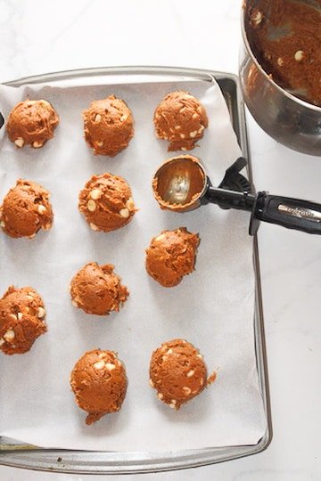 Overhead photo of a parchment-lined baking sheet with multiple rounded scoops of dough. A metal mixing bowl with remaining dough sits in the top right, and a cookie scoop rests on the tray, showing the step of portioning chilled dough into even mounds before baking.