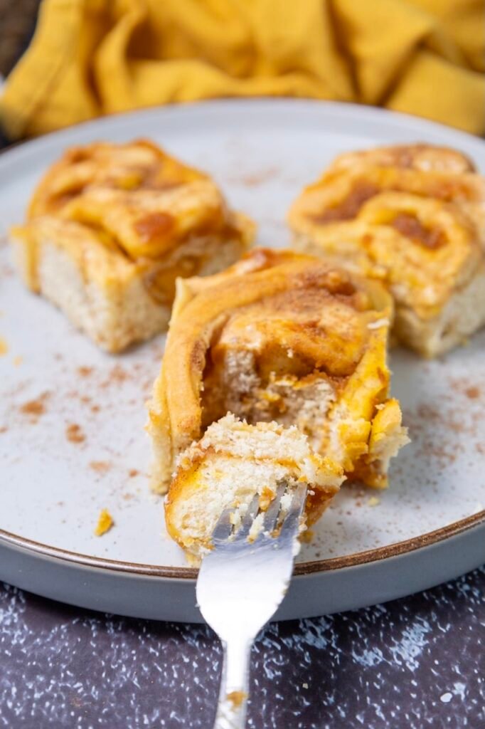 A close-up of soft pumpkin cinnamon rolls with cream cheese frosting, showing a fork cutting into the fluffy interior with visible pumpkin filling and cinnamon swirl on a plate.
