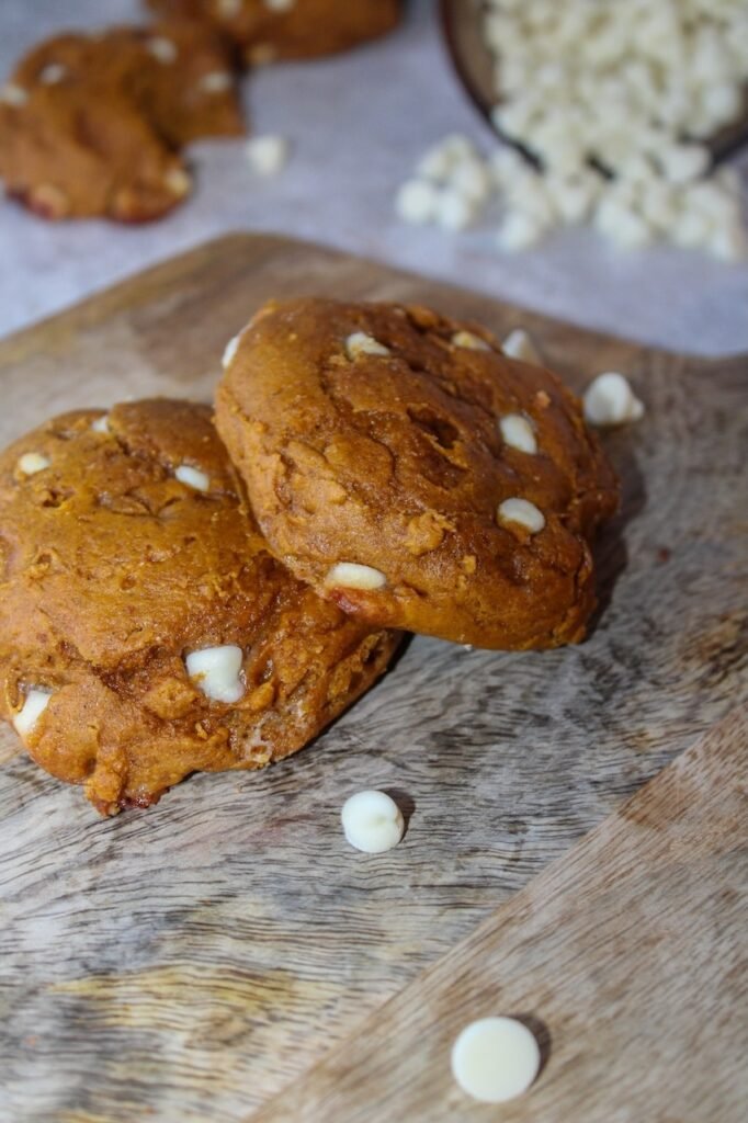 Two thick, soft pumpkin spice cookies studded with white cream cheese-flavored baking chips sit on a rustic wooden board. A few baking chips are scattered in the foreground, and more cookies and a bowl of chips appear slightly blurred in the background, highlighting the cookies’ pillowy texture and mix-ins.