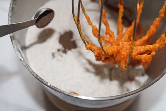 Overhead view inside a metal stand mixer bowl with pale spice cake mix added on top of the pumpkin mixture. A whisk attachment is lifted above the batter with orange pumpkin clinging to the wires, and a measuring spoon with ground spice sits beside the bowl, showing the step of adding the dry cake mix before mixing into cookie dough.