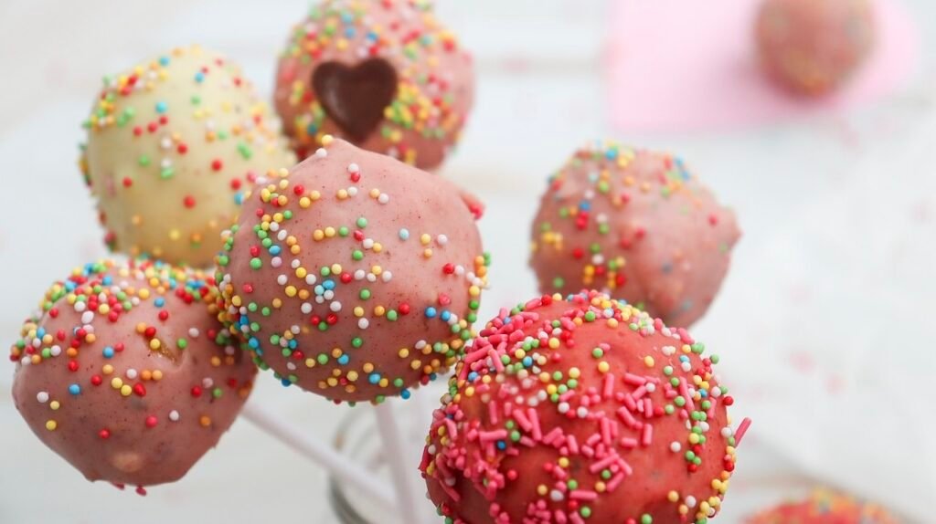 Close-up view of colorful Valentine cake pops coated in pink and white chocolate and covered in bright sprinkles, perfect for holiday treat boards and party desserts.