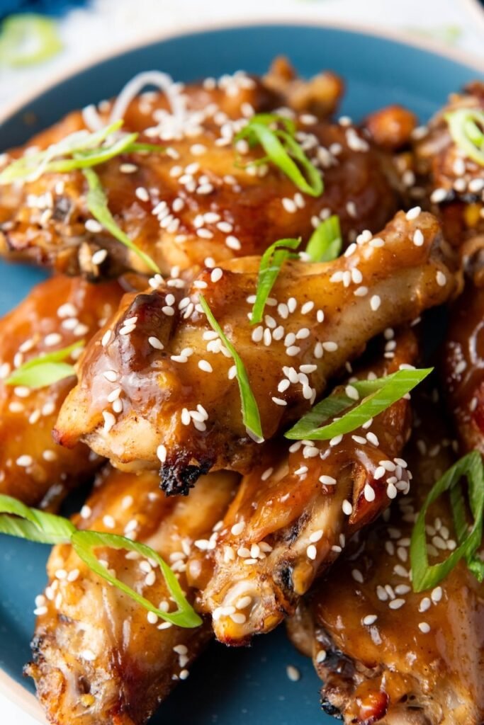Close-up of glazed Asian sticky chicken wings piled on a blue plate, coated in a shiny brown sauce and topped with toasted sesame seeds and thinly sliced green onions.