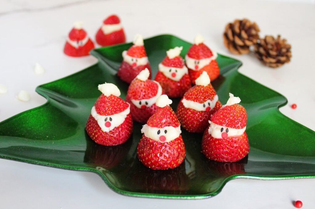 Overhead view of Strawberry Santas arranged on a green Christmas tree–shaped serving platter, surrounded by scattered white chocolate chips, red sprinkles, and two pinecones on a marble surface, creating a festive holiday presentation.