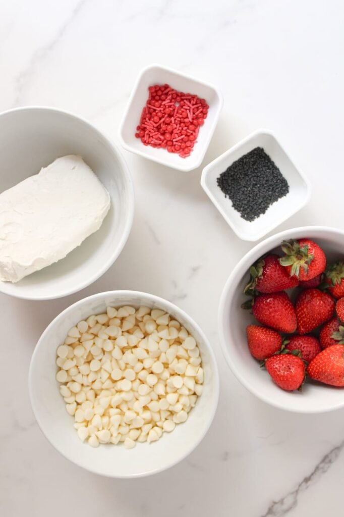 Ingredients for Strawberry Santas arranged in bowls, including strawberries, white chocolate chips, cream cheese, red sprinkles, and black sesame seeds.