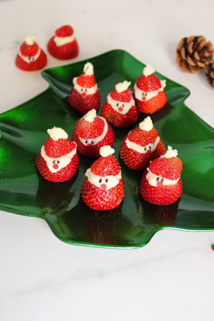 A group of finished Strawberry Santas arranged on a green tree-shaped serving platter, each made from a strawberry base filled with white chocolate cream cheese and topped with a small strawberry “hat,” decorated with sprinkle eyes and noses for a festive holiday presentation.