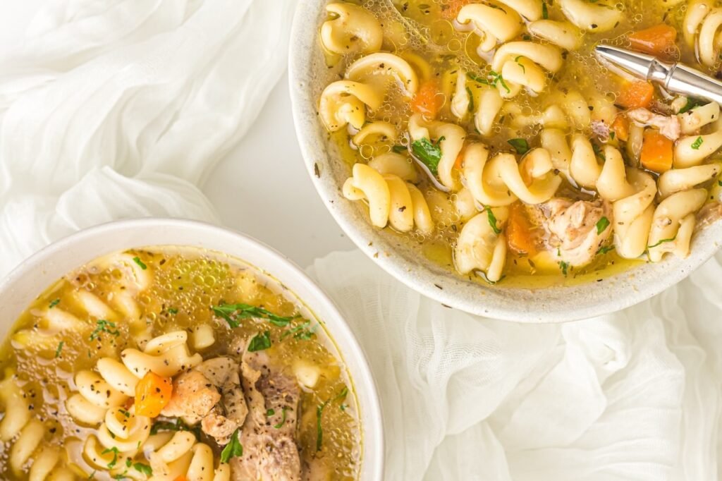 Overhead photo of two bowls of chicken noodle soup filled with spiral noodles, carrots, celery, chicken, and herbs, placed on a white cloth.