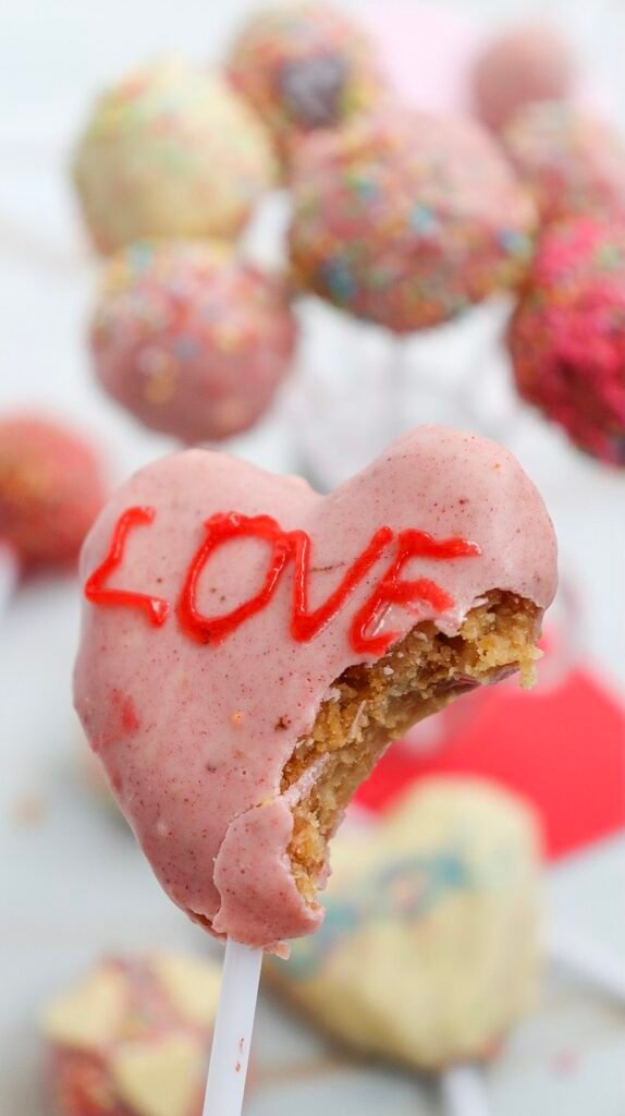 Close-up shot of a heart-shaped Valentine cake pop with a bite taken out, showing the soft cake center and a LOVE design on top.