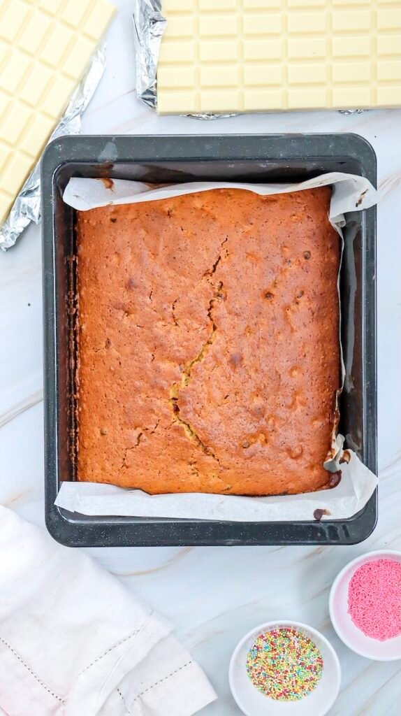 Overhead photo of a golden baked cake in a lined pan with sprinkles and white chocolate nearby, the cooling step before making Valentine’s Day cake pops.