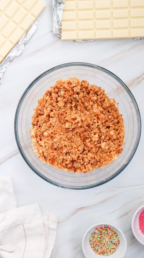 Overhead view of a bowl of finely crumbled cake surrounded by white chocolate and sprinkles, the step before mixing and shaping Valentine’s Day cake pops.