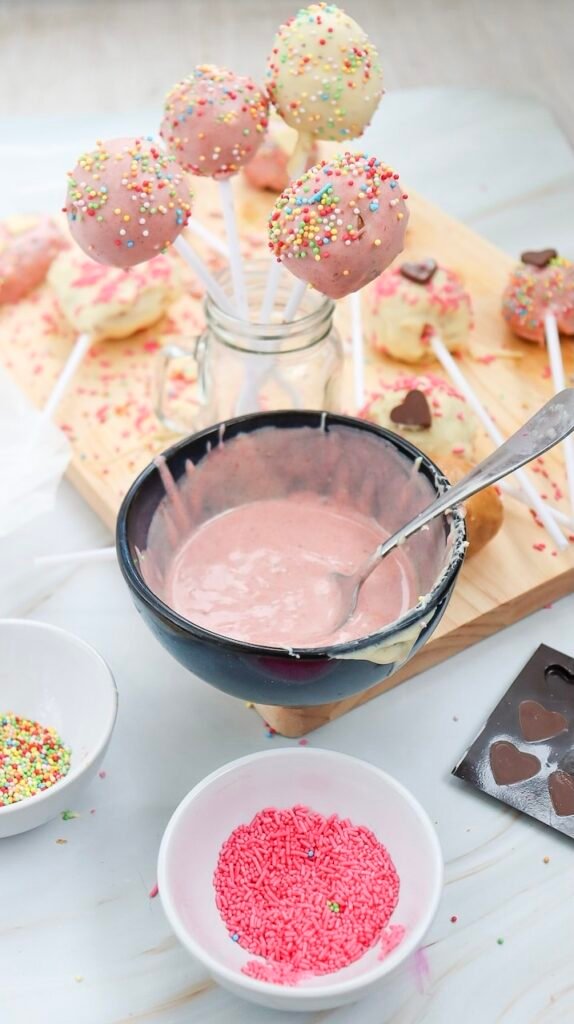 Bright Valentine’s Day cake pops coated in pink and white chocolate with rainbow sprinkles, shown drying upright while a bowl of pink melted chocolate sits in front.