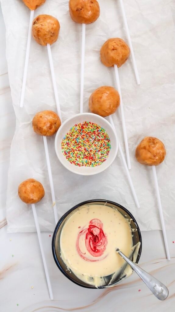 Overhead photo of undipped cake pops on sticks with a bowl of white chocolate swirled with pink coloring and rainbow sprinkles, the decorating setup for Valentine’s Day cake pops.
