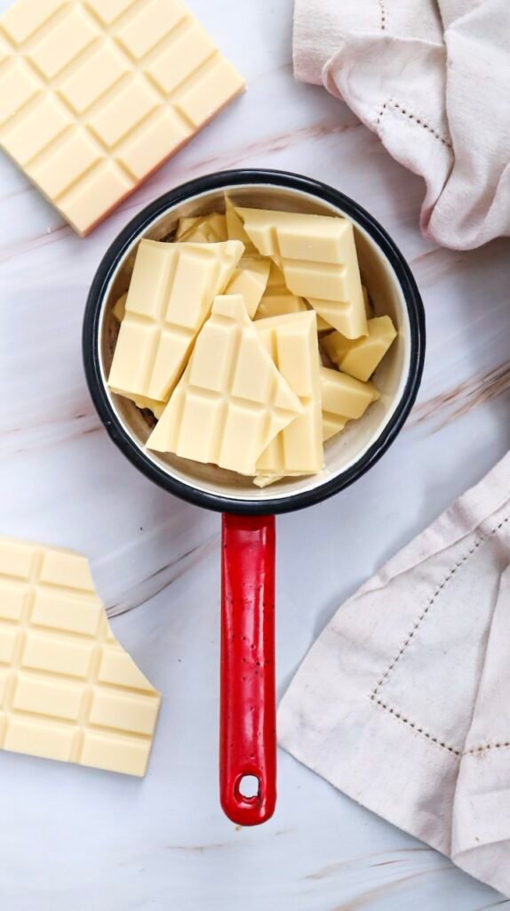 Overhead view of broken white chocolate pieces in a red-handled saucepan, the melting step for smooth Valentine’s Day cake pop coating.