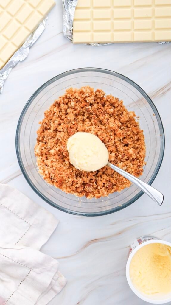 Overhead photo of frosting being added to a bowl of crumbled cake with white chocolate nearby, the mixing step for soft and rollable Valentine’s Day cake pops.