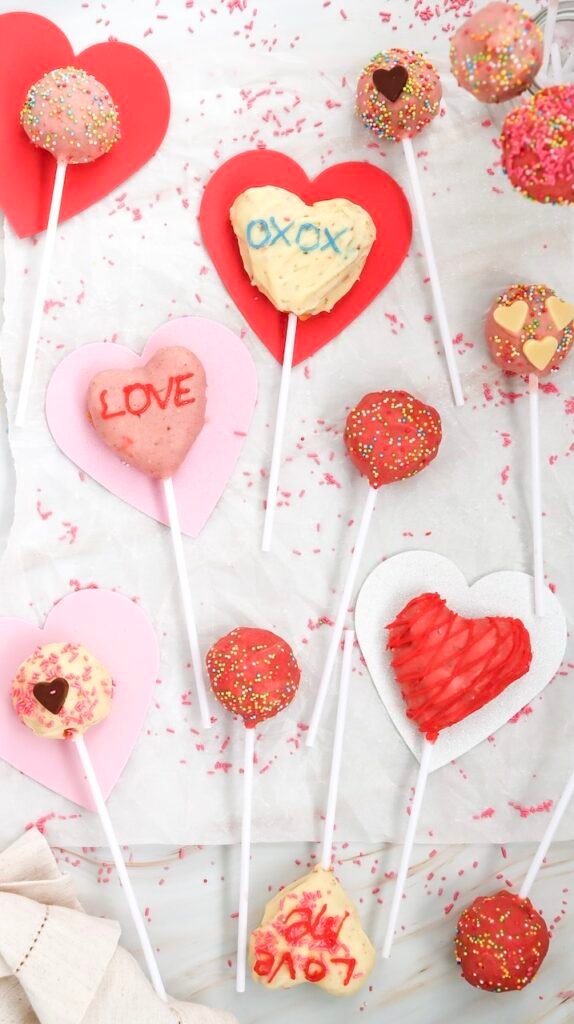 Overhead photo of finished Valentine’s Day cake pops decorated with heart shapes, sprinkles, and handwritten messages on colorful heart backdrops.