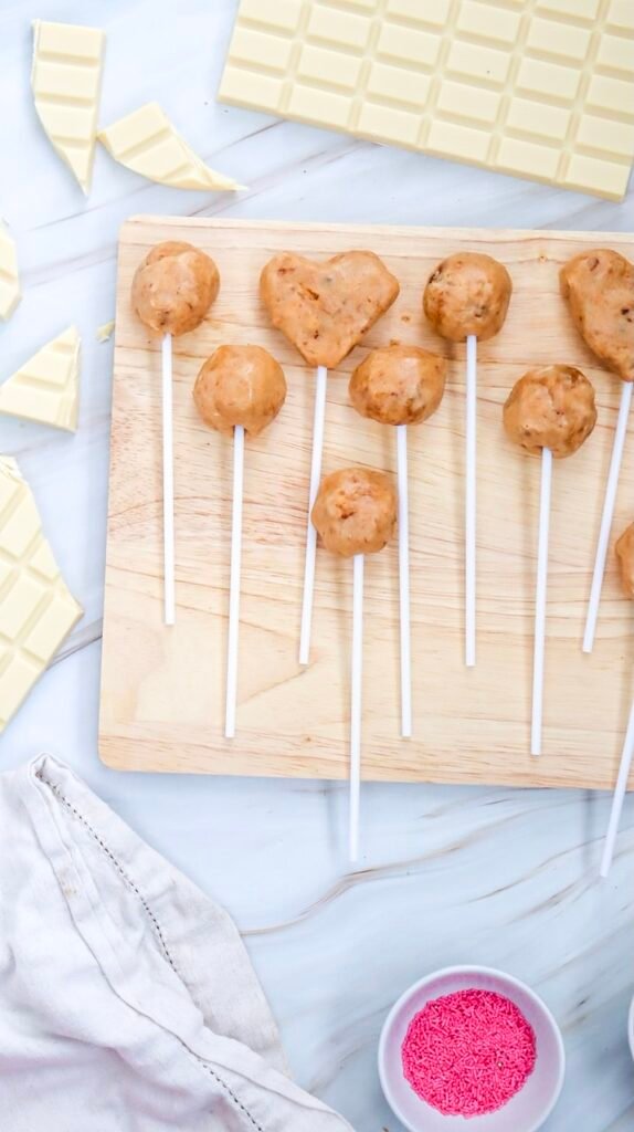 Overhead view of round and heart-shaped cake pops on sticks placed on a wooden board with white chocolate pieces nearby, the shaping step for Valentine’s Day cake pops.