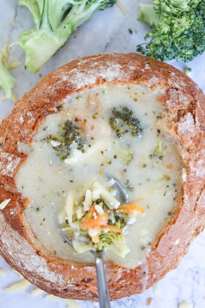 A spoon lifting a scoop of creamy broccoli cheddar soup from a round bread bowl, showing tender broccoli florets, soft potato pieces, and shredded cheddar melted into the soup. The soup looks thick and smooth, with visible vegetables and cheese, sitting on a marble surface with extra broccoli nearby, indicating a hearty, ready-to-eat comfort dish.