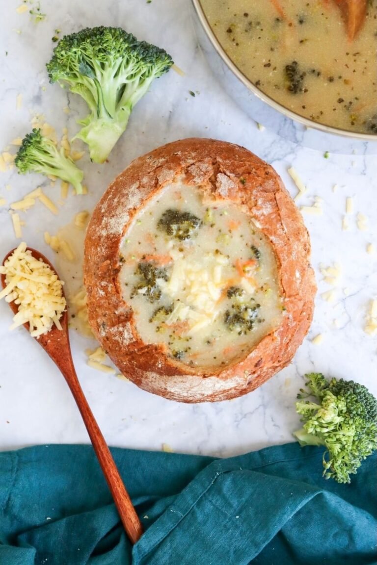 A round bread bowl filled with creamy broccoli cheddar soup, topped with shredded cheddar cheese, sitting on a marble surface. Pieces of broccoli and carrot are visible in the thick soup, with extra broccoli florets, shredded cheese, and a wooden spoon nearby, showing the soup ready to serve as a comforting main dish.