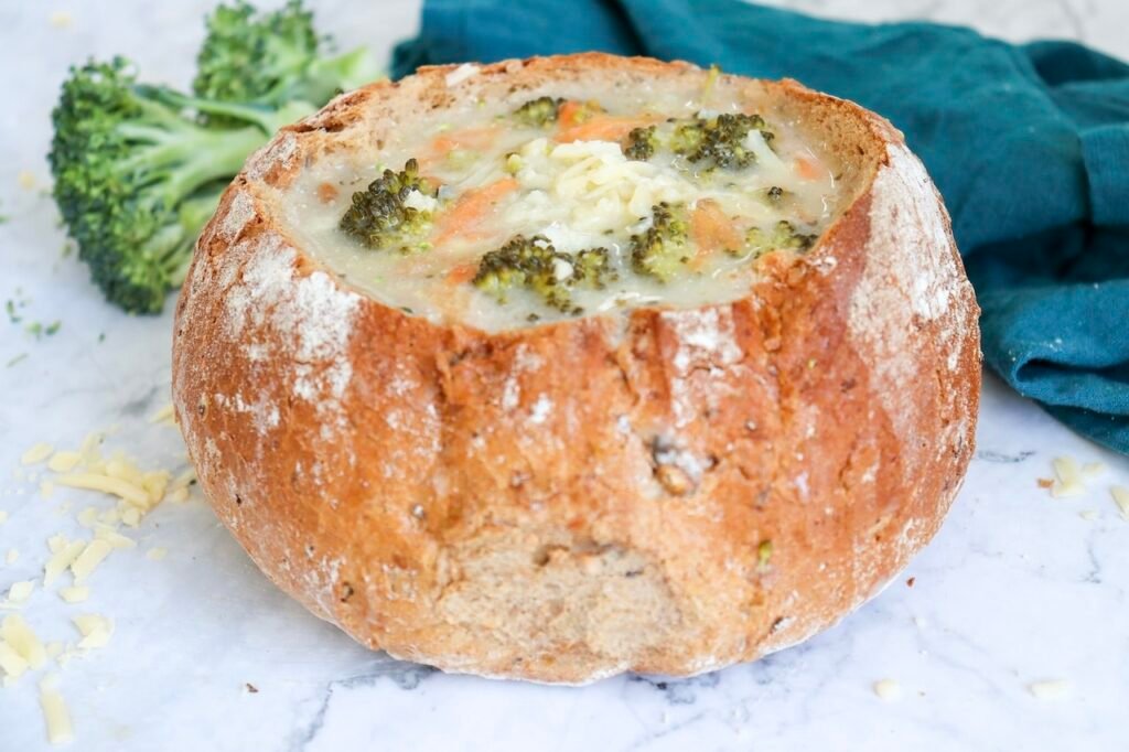 A round loaf of bread hollowed out and filled with creamy broccoli cheddar soup, showing pieces of broccoli and carrot in a thick, pale soup. The bread bowl sits on a marble surface with extra broccoli florets and shredded cheese nearby, indicating the soup is ready to serve as a warm, comforting meal.