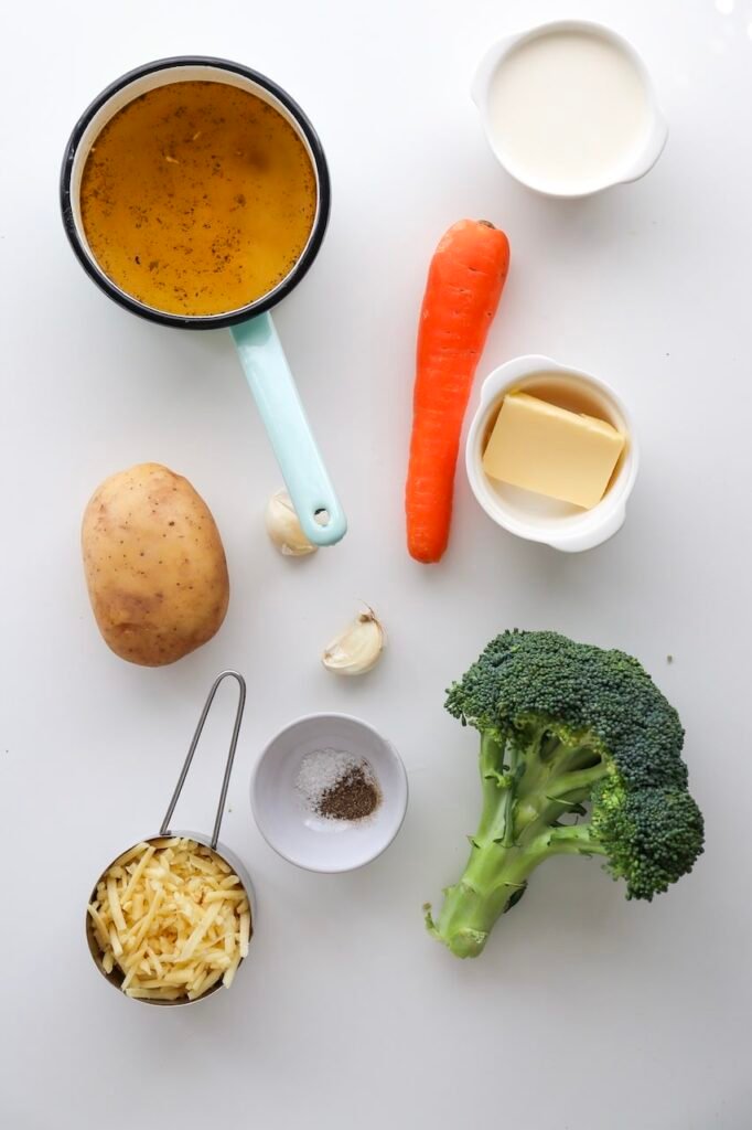 Overhead view of broccoli cheddar soup ingredients laid out on a white surface, including fresh broccoli, shredded cheddar cheese, potato, carrot, garlic, butter, cream, broth, salt, and pepper.