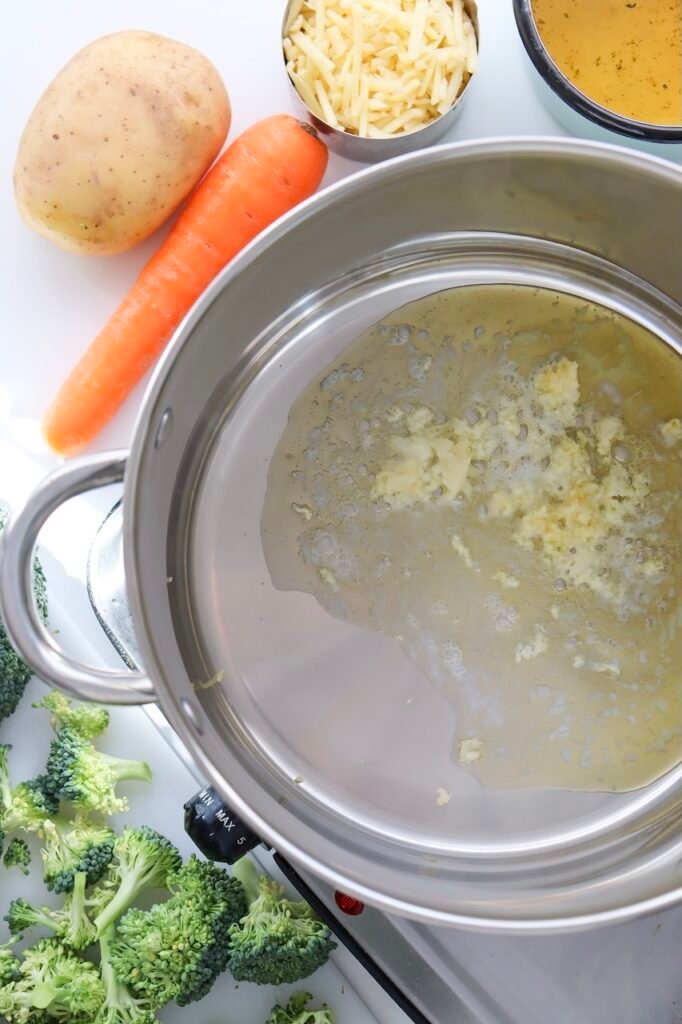 Butter melting with minced garlic in a stainless steel pot on the stovetop, beginning the base , with carrot, potato, cheese, and broth visible nearby.