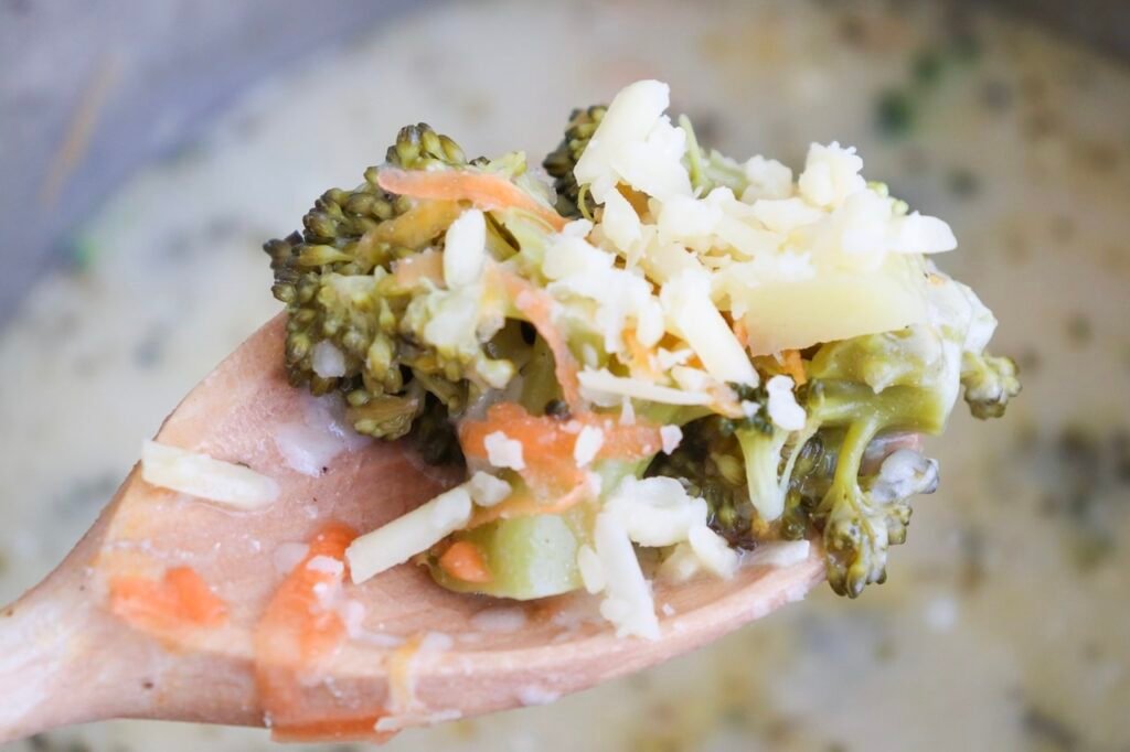 A close-up view of a wooden spoon lifting a scoop of broccoli cheddar soup, showing tender broccoli florets, soft carrot shreds, and melted cheddar cheese coated in a thick, creamy base. The soup appears smooth and rich without being heavy, with visible vegetables indicating it is fully cooked and ready to serve.