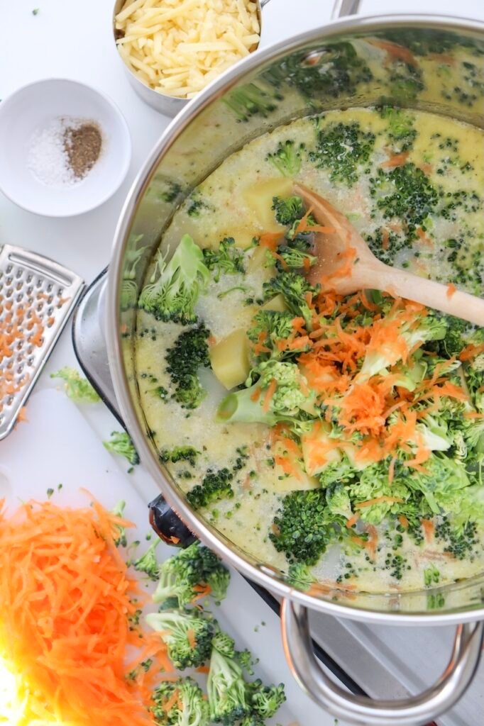 florets, diced potato, and grated carrot simmering in a base inside a stainless steel pot on the stovetop. The soup looks pale and lightly bubbling around the edges, showing gentle heat, with shredded  cheese, salt, pepper, and extra vegetables visible nearby on a white surface.