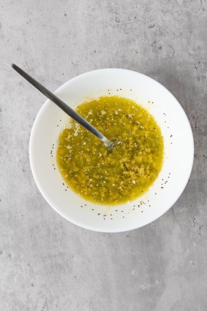 Overhead view of a bowl of melted butter mixed with Dijon mustard, minced onion, and poppy seeds with a spoon, prepared as a glaze.
