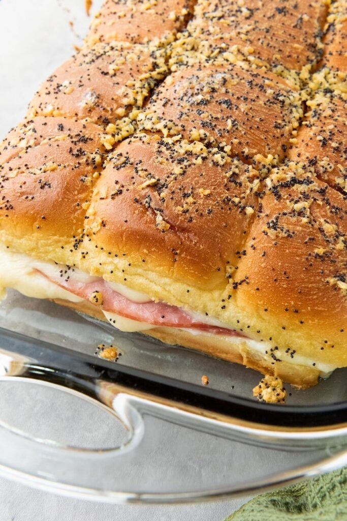 Close-up of baked ham and cheese sliders in a glass baking dish, showing golden buns topped with poppy seeds and a buttery glaze, with layers of ham and melted cheese visible along the side.