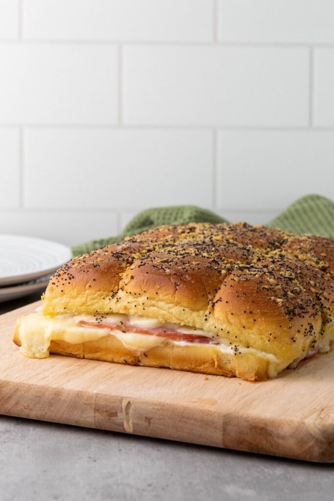 Baked ham and cheese sliders on a wooden cutting board, showing soft golden buns topped with poppy seeds and layers of melted cheese and ham along the cut edge, ready to serve.