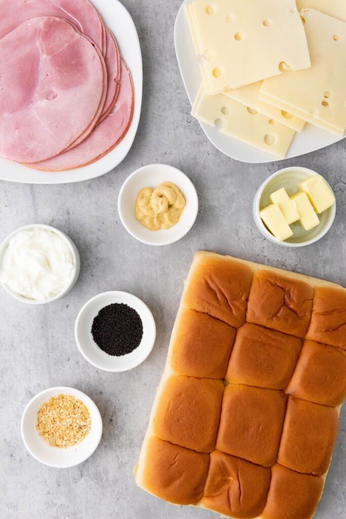 Flat lay of ham and cheese slider ingredients including soft slider buns, sliced ham, Swiss cheese, mayonnaise, Dijon mustard, butter, poppy seeds, and minced onion arranged on a gray surface.