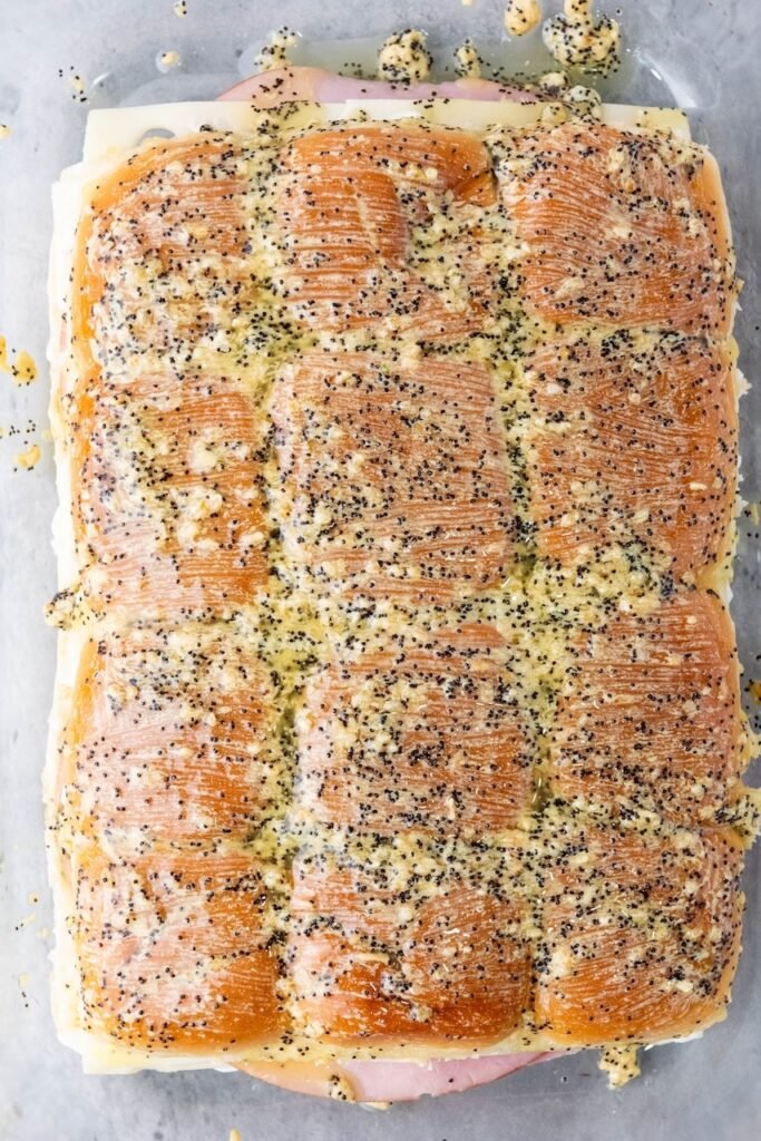 Overhead view of assembled sanwhiches brushed with a buttery poppy seed glaze, arranged in a baking dish just before going into the oven.
