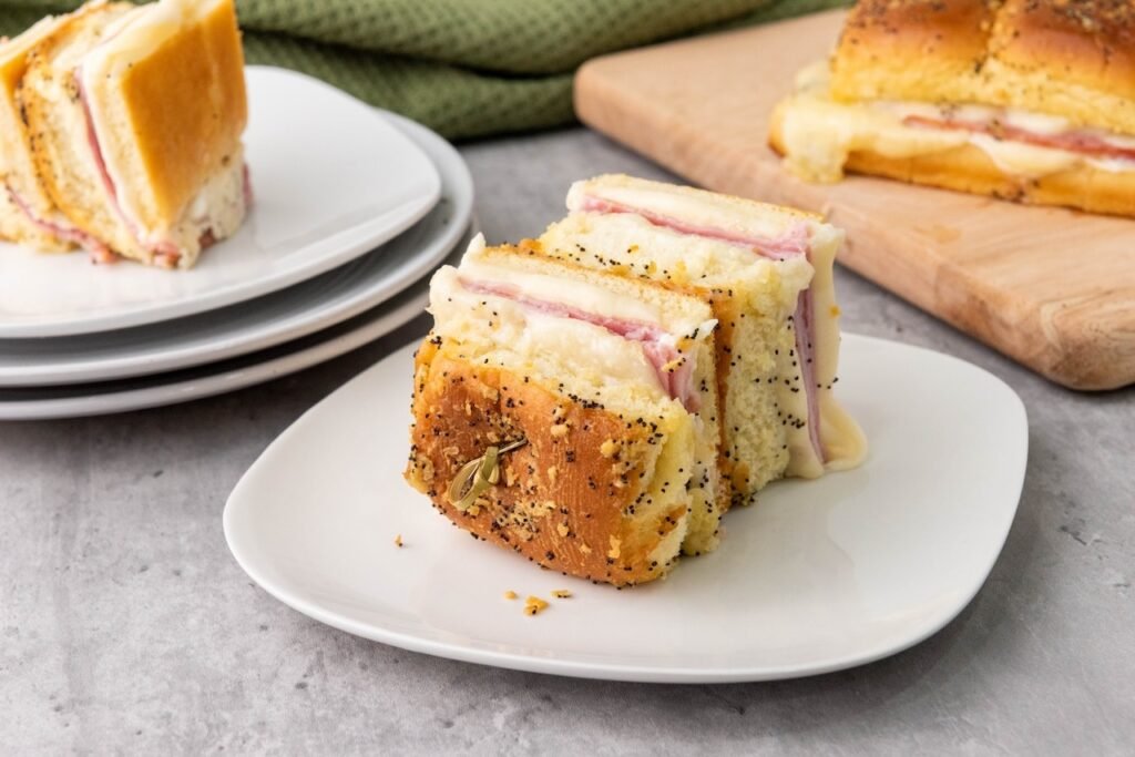 A single ham and cheese slider on a white plate, showing soft golden buns layered with sliced ham and melted cheese, lightly coated in poppy seed glaze and served warm and ready to eat.