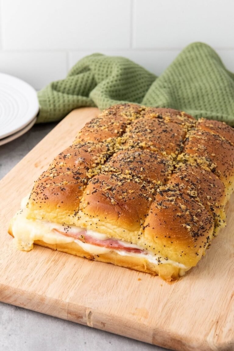 Pull-apart ham and cheese sliders on a wooden cutting board with one slider removed, showing melted cheese and ham inside and golden poppy seed tops.