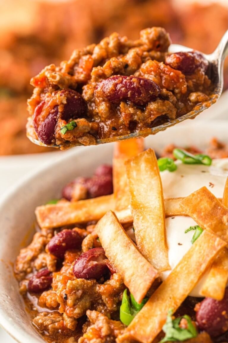 A close-up of a spoonful of thick Instant Pot chili with ground beef and kidney beans held above a bowl of chili topped with sour cream, green onions, and crispy tortilla strips.